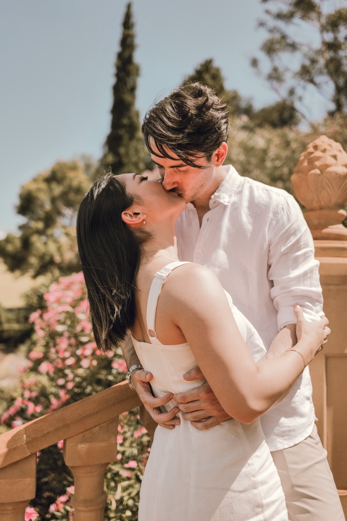 Damon kissing Christina by a sandstone balustrade with pink flowers behind
