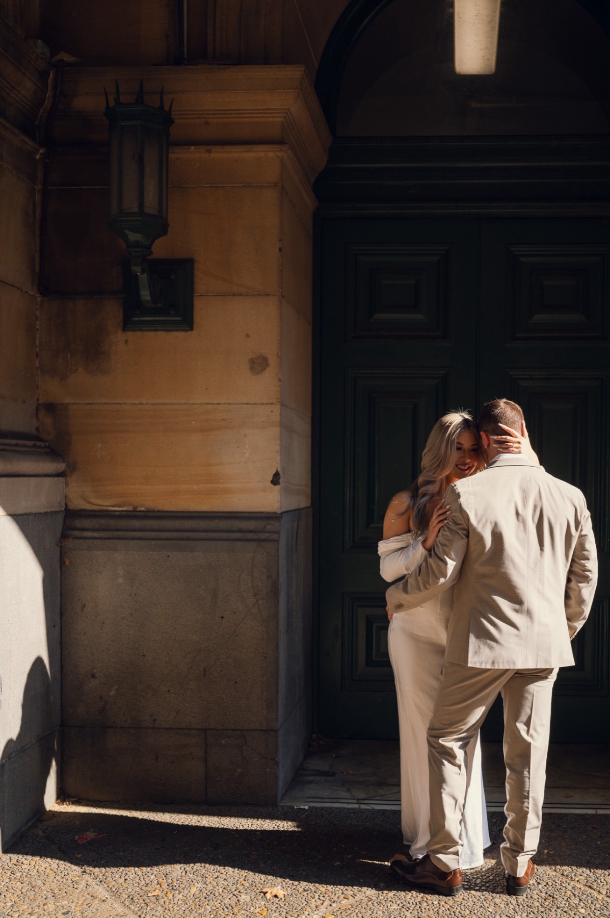 Dave and Kim embracing in a patch of sunlight beside a tall dark doorway in a sandstone building