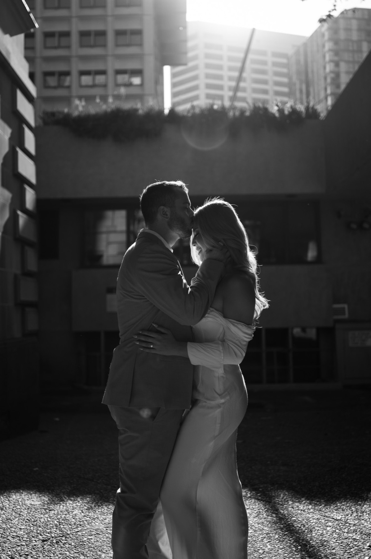 Black and white photo of the couple kissing, backlit by sun between city buildings