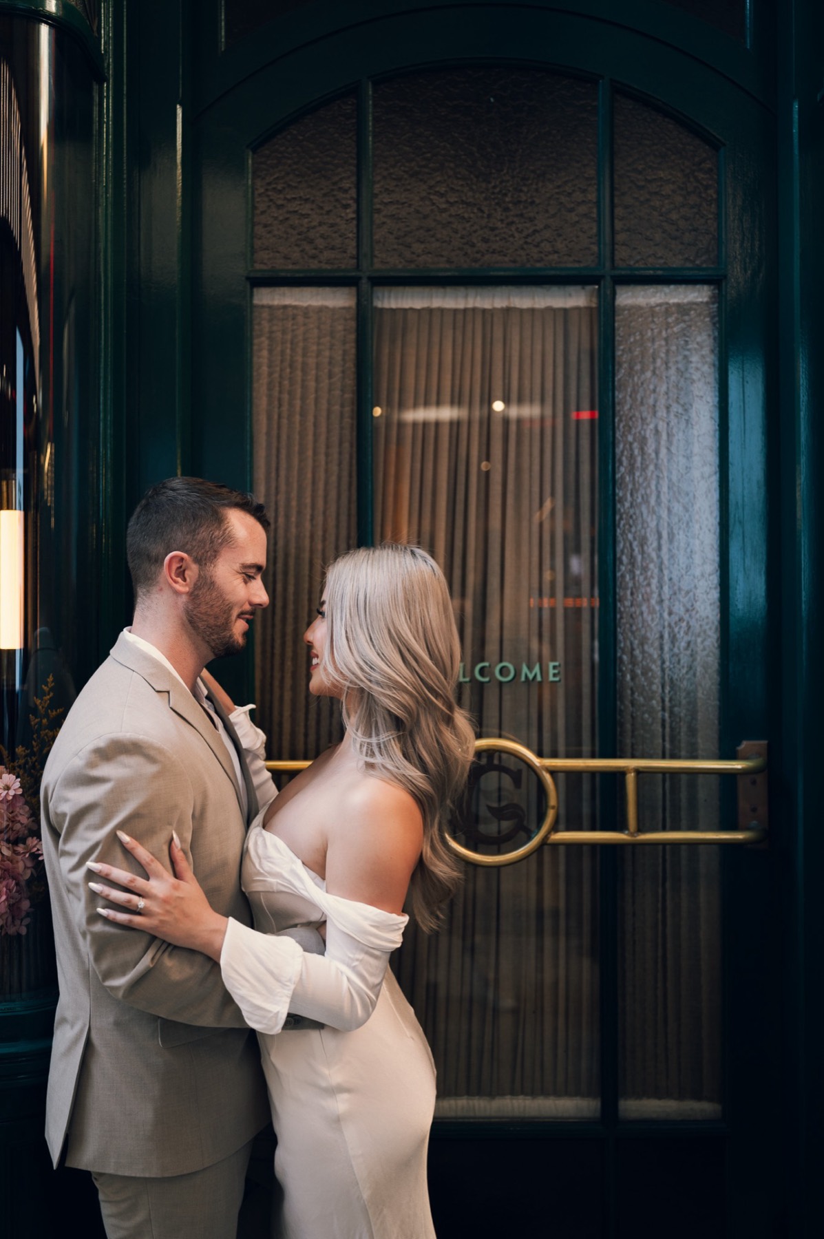 Dave and Kim holding each other at a brass-trimmed hotel entrance with a Welcome sign
