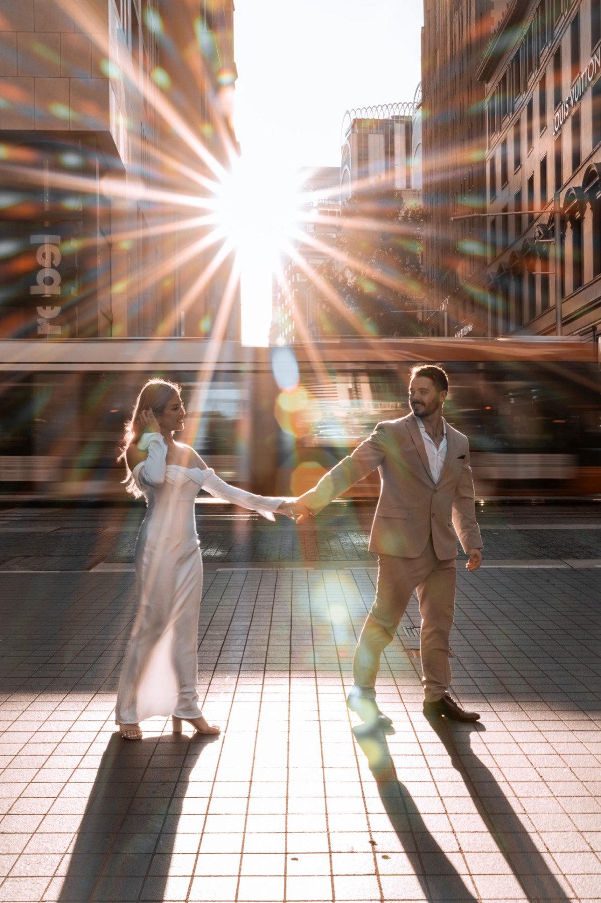 The couple holding hands across a city street as the sun bursts between buildings