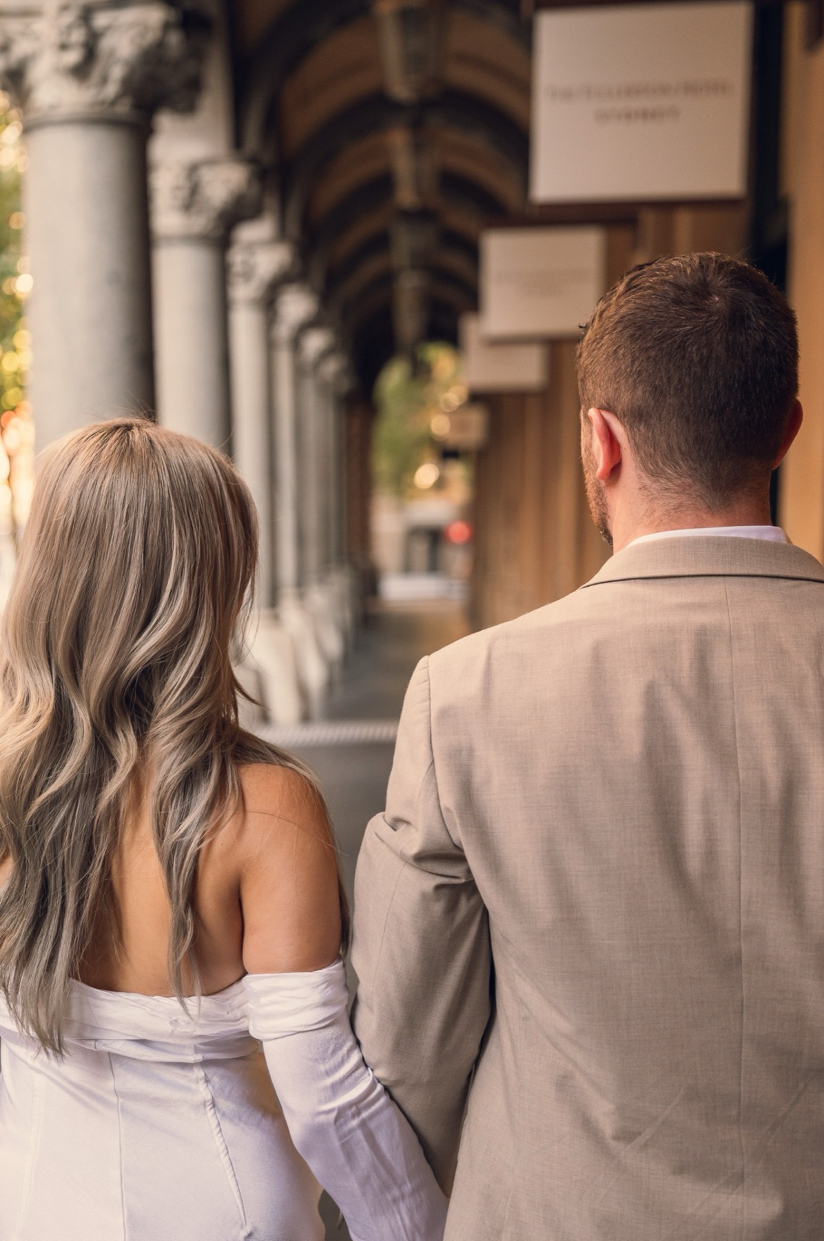 Dave and Kim walking hand in hand down a sandstone colonnade, seen from behind