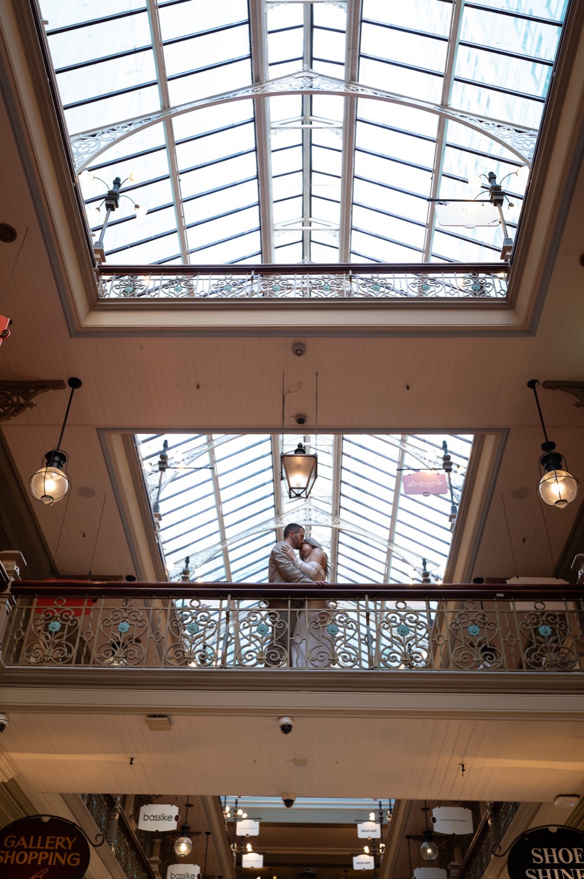 The couple kissing on an upper balcony beneath an arched glass arcade roof