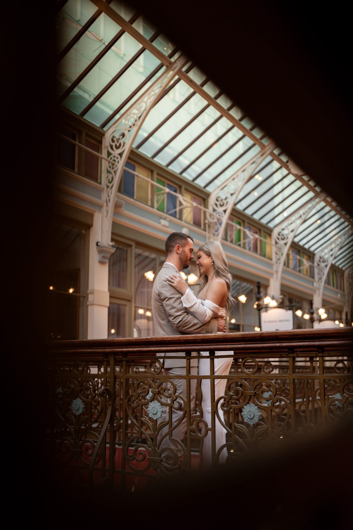 Dave and Kim embracing at an ornate iron balcony railing under a glass roof