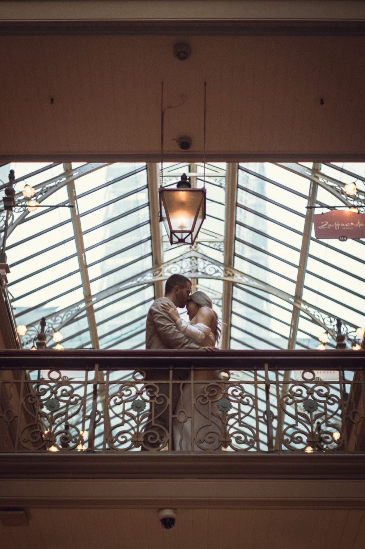 Dave and Kim embracing on a balcony framed by an arcade's glass ceiling and lantern