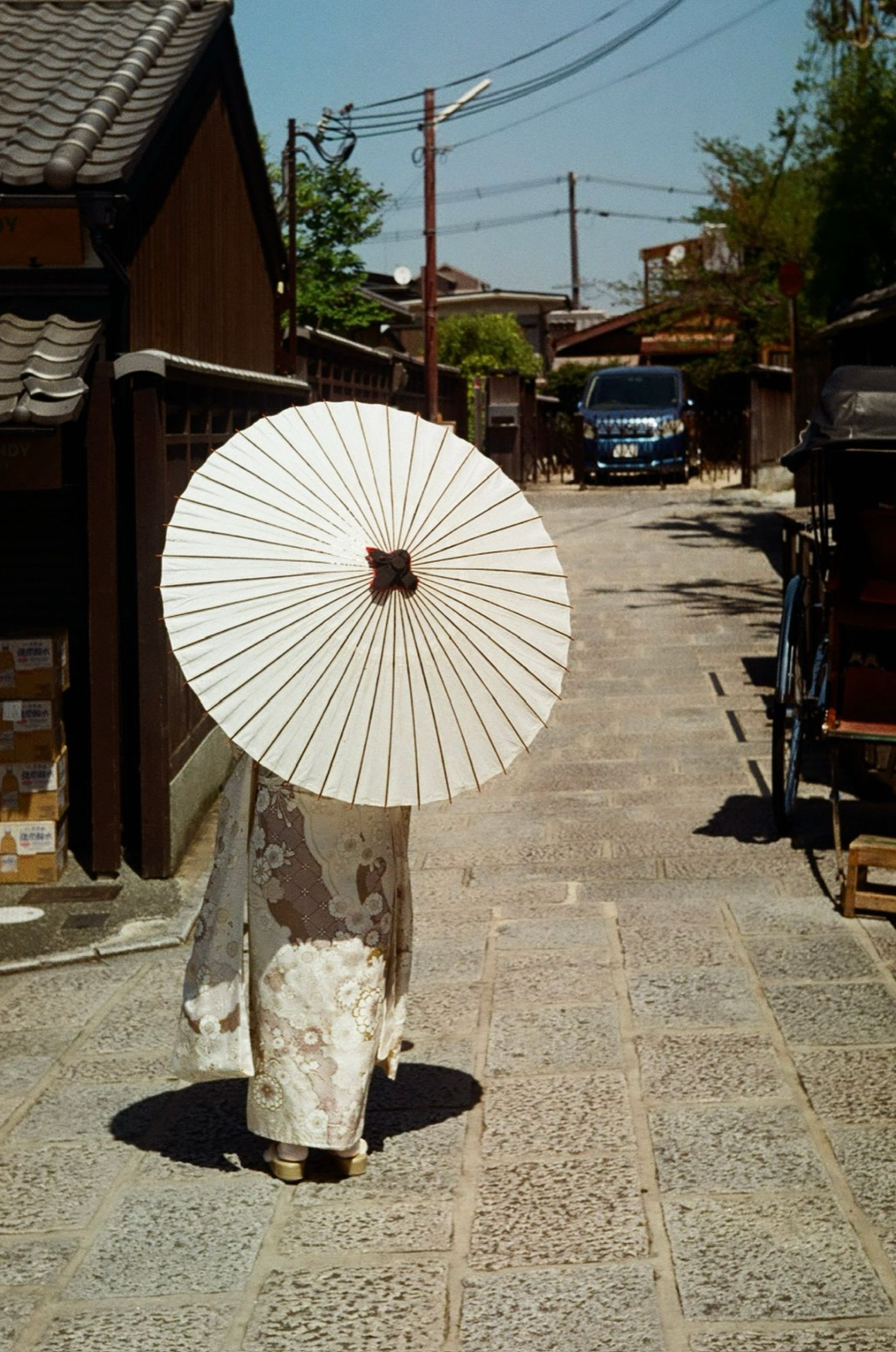 A woman in a floral kimono walks a sunlit stone lane beneath a white paper parasol