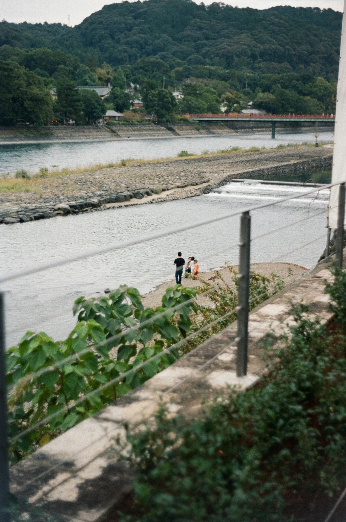 Two figures on a riverbank below green hills, seen through a railing