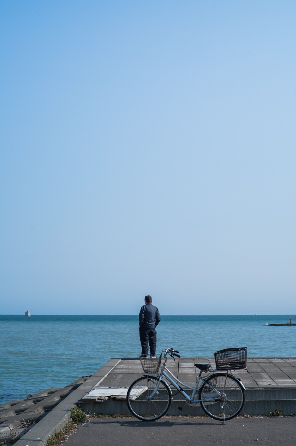 A man looks out over a calm blue sea, his bicycle parked on the sea wall behind him