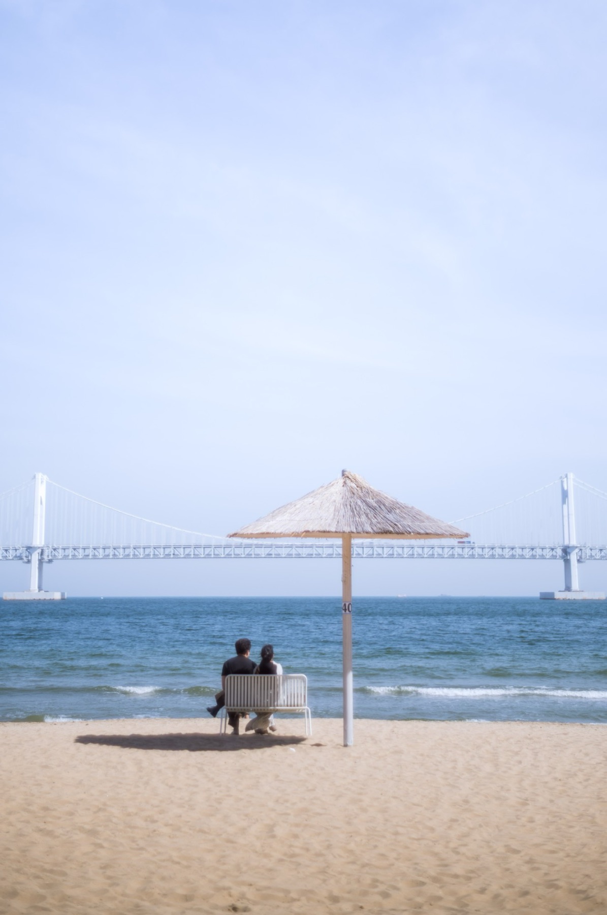 A couple on a beach bench beneath a thatched umbrella, Gwangan Bridge stretched across the bay