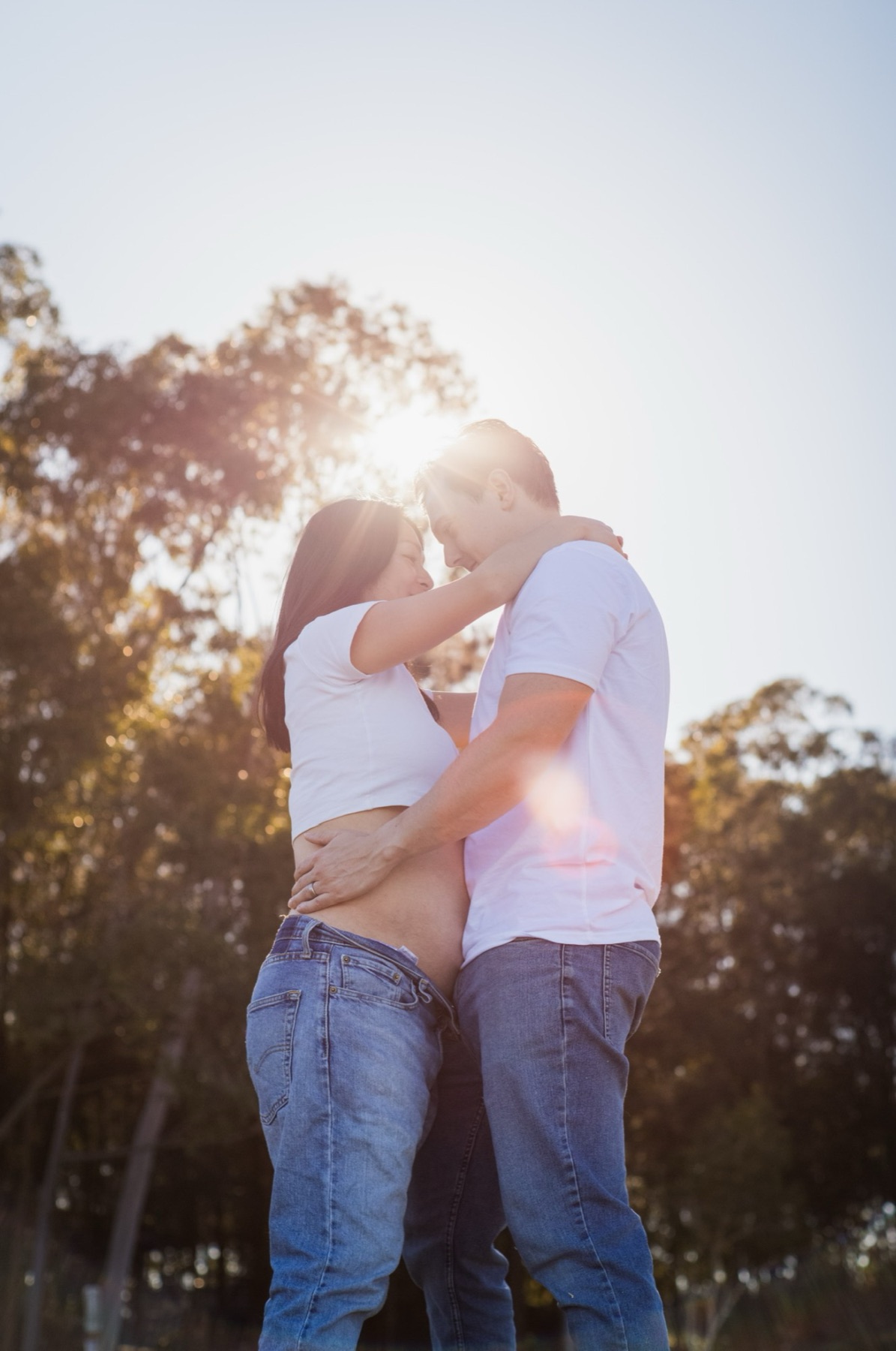An expecting couple in white T-shirts and jeans embracing under eucalyptus trees, the sun flaring between them