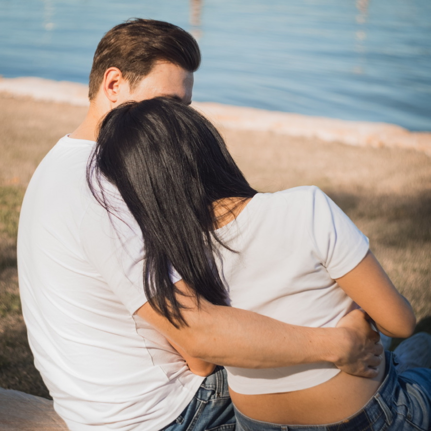 The couple seen from behind sitting by the water, her head resting on his shoulder and his arm around her bump