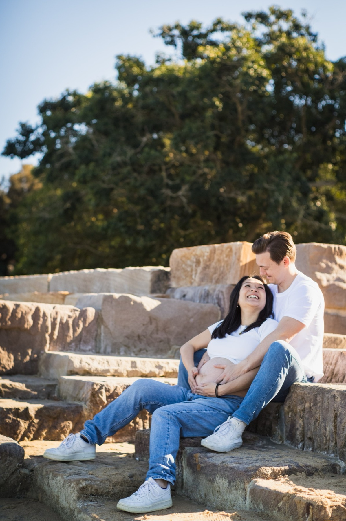 The mother-to-be laughing up at her partner as they sit together on sunlit sandstone steps, hands on her bump
