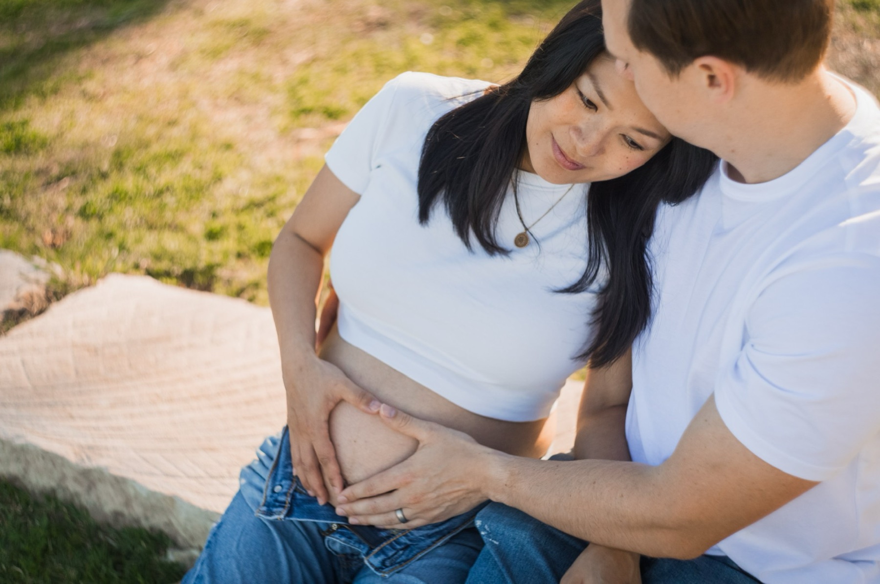 Close-up from above of the couple sitting on the grass, their hands making a heart shape on her bump
