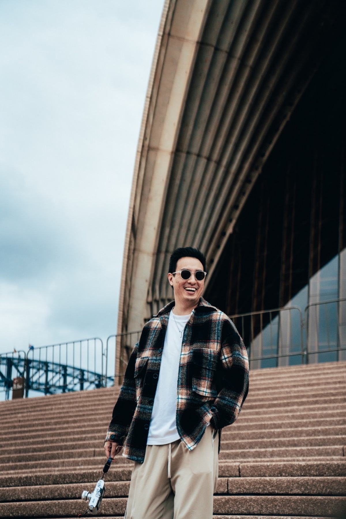 Ryan Chong, the photographer behind thirtysixframes, laughing on the Sydney Opera House steps in a flannel shirt and sunglasses, a film camera in hand