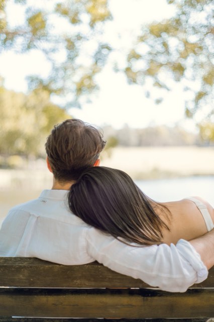 Damon and Christina seen from behind on a park bench by a lake, her head resting on his shoulder
