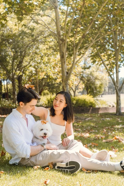 The couple sitting on the grass in a sunny park with their fluffy white dog, Marlo