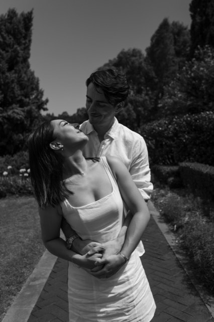 Black and white photo of Damon holding Christina from behind on a garden path, both smiling