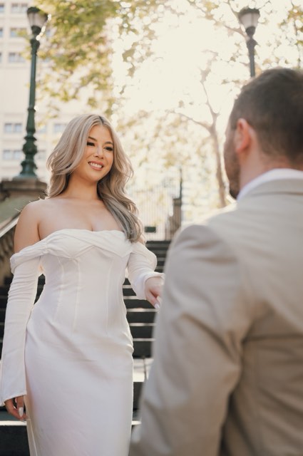 Kim in an off-the-shoulder white dress smiling at Dave on the stairs, trees and lamp posts behind