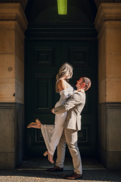 Dave lifting Kim off the ground in front of a heavy dark door, her shoes kicked up behind her