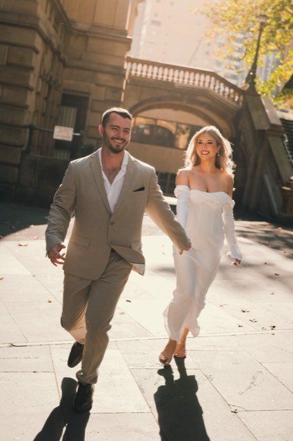 Dave and Kim running hand in hand across a sunny square, laughing, sandstone archway behind
