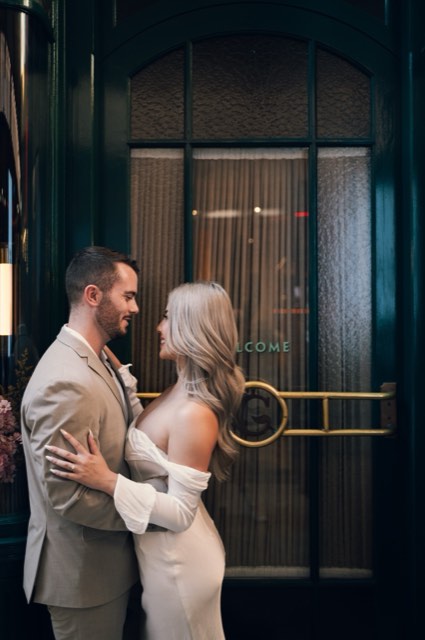 Dave and Kim holding each other at a brass-trimmed hotel entrance with a Welcome sign
