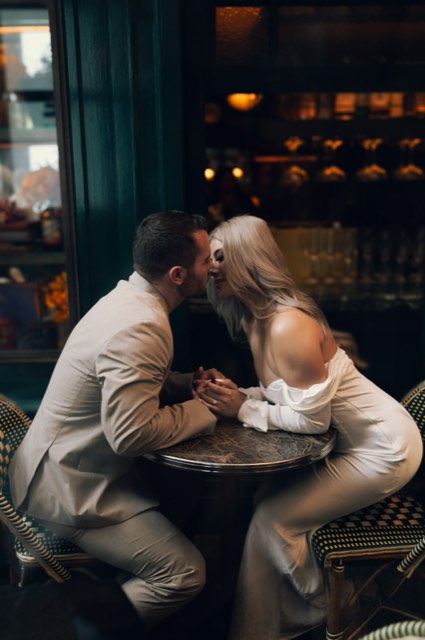 The couple leaning in for a kiss across a small marble café table, holding hands