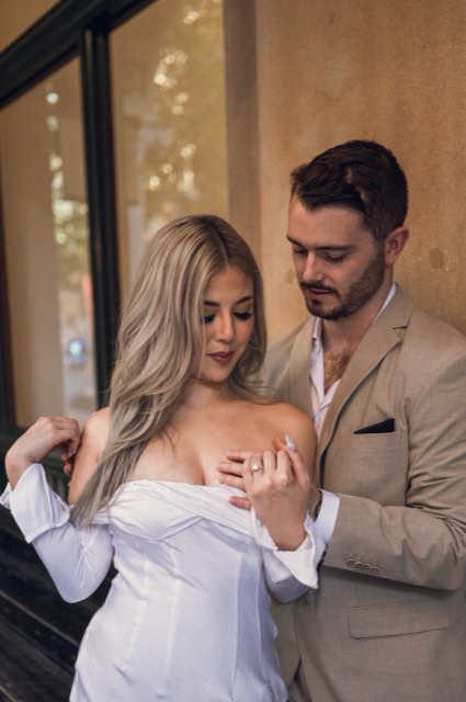 Kim resting her hand on her chest as Dave holds her from behind by a warm-lit window