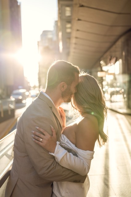 Dave kissing Kim's forehead on a sunlit city street, her hand on his back